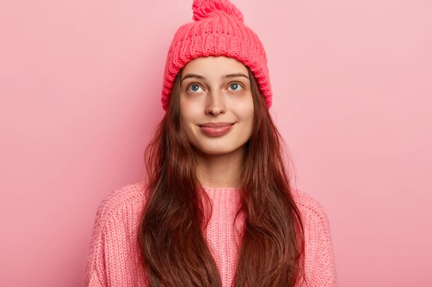 Una ragazza millenaria bruna soddisfatta e sognante guarda sopra con pensieri piacevoli, indossa un cappello e un maglione lavorato a maglia vivaci, modelli su sfondo rosa, ha un sorriso affascinante e un aspetto tenero