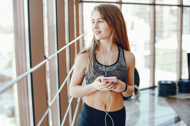 Una ragazza sportiva bella e atletica si leva in piedi vicino alla finestra in palestra con il telefono