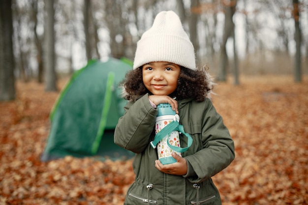 Una ragazzina nera in tenda da campeggio nella foresta Ragazza tiene in mano un thermos Ragazza nera che indossa un cappotto color cachi e un cappello beige