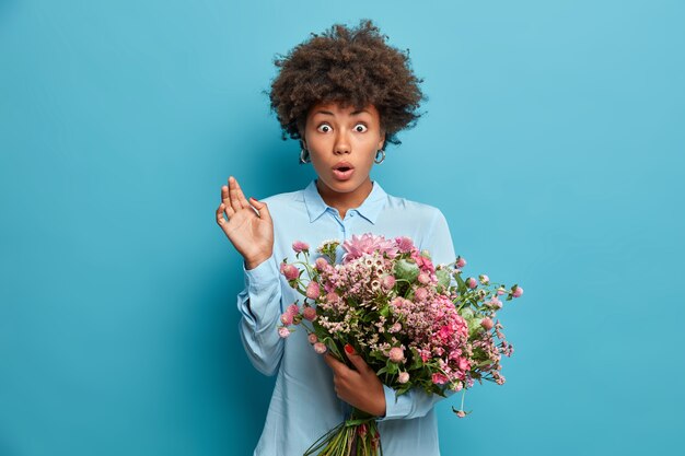 Una signora afroamericana dai capelli ricci e scioccata riceve un mazzo di fiori da una persona sconosciuta, fissa gli occhi spalancati mentre riceve una consegna inaspettata, indossa un'elegante camicia blu, si trova al coperto. Concetto floreale