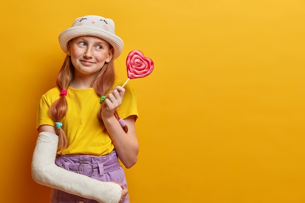 Una sognante ragazza lentigginosa positiva posa con un grande lecca-lecca a forma di cuore, ha un debole per i dolci, ama mangiare cibi nocivi, detiene deliziose caramelle, indossa abiti estivi alla moda, ha un braccio rotto. Dipendenza da zucchero