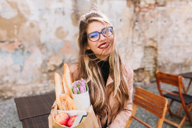 Una splendida ragazza dopo il servizio fotografico ha acquistato cibo fresco e beve caffè godendosi una giornata di sole. Elegante giovane fotografo femminile che tiene il sacchetto della spesa e la tazza di cappuccino in posa nella caffetteria all'aperto.