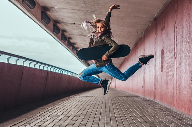 Una studentessa felice con i capelli biondi, vestita con abiti alla moda, salta con uno skateboard sul marciapiede del ponte.