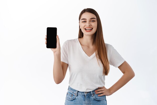 Una studentessa sorridente mostra l'app per lo shopping, lo schermo dello smartphone vuoto e sembra soddisfatta, consiglia un negozio o un'applicazione online, si trova su uno sfondo bianco