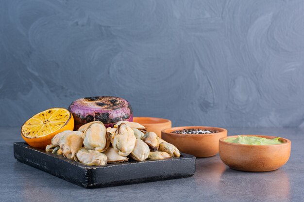 Una tavola di legno nera di conchiglie cotte con cipolla fritta e limone a fette su una superficie di pietra