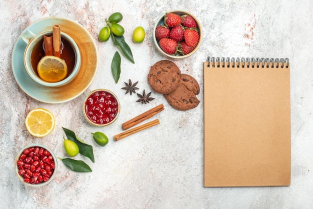 una tazza di tè ciotole di frutti di bosco agrumi anice stellato e bastoncini di cannella biscotti al cioccolato accanto al quaderno di crema la tazza di tè con cannella sul tavolo