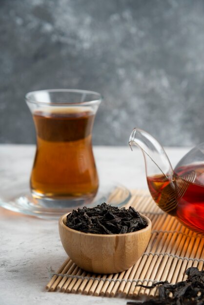 Una tazza di tè di vetro con tè sciolti secchi e teiera.