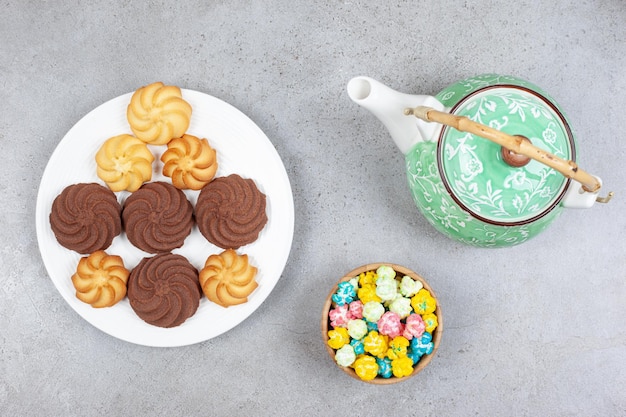 Una teiera decorata, una ciotola di caramelle e un piatto di biscotti sulla superficie di marmo