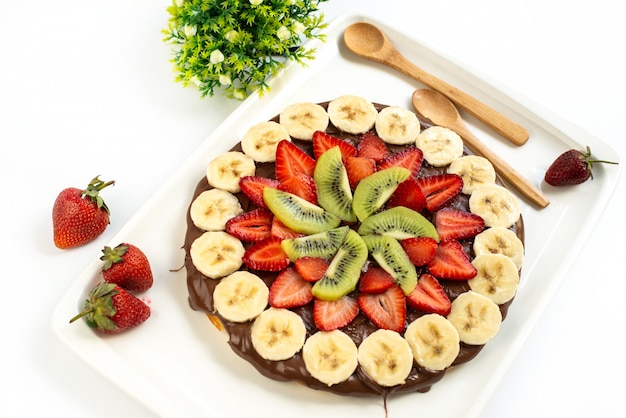 Una torta di frutta al cioccolato vista dall'alto deliziosa con frutta a fette insieme a fragole e cucchiai di legno all'interno di frutta zucchero biscotto scrivania bianca