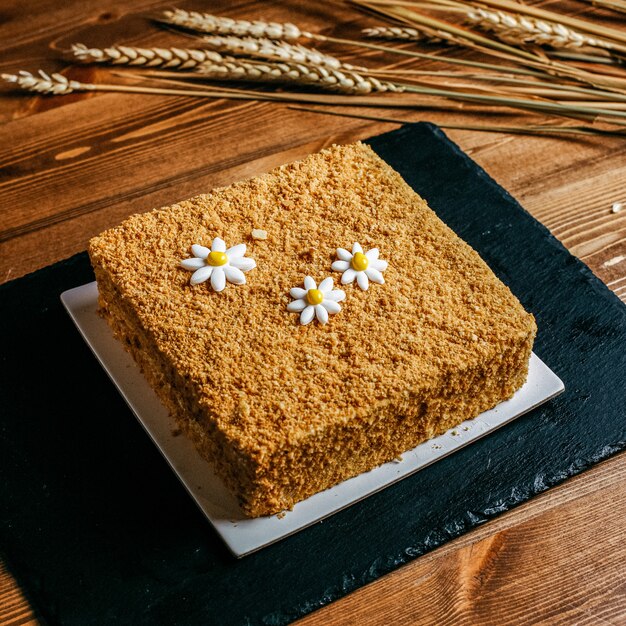 Una torta di miele di vista frontale decorata con il quadrato della camomilla ha formato la torta di compleanno deliziosa dentro il compleanno bianco della dolcezza della confetteria del piatto sui precedenti marroni