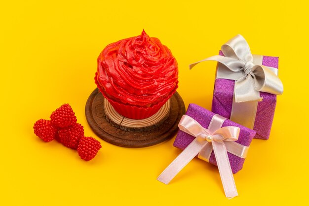 Una torta rossa vista frontale con lamponi freschi e contenitori di regalo viola sullo scrittorio giallo