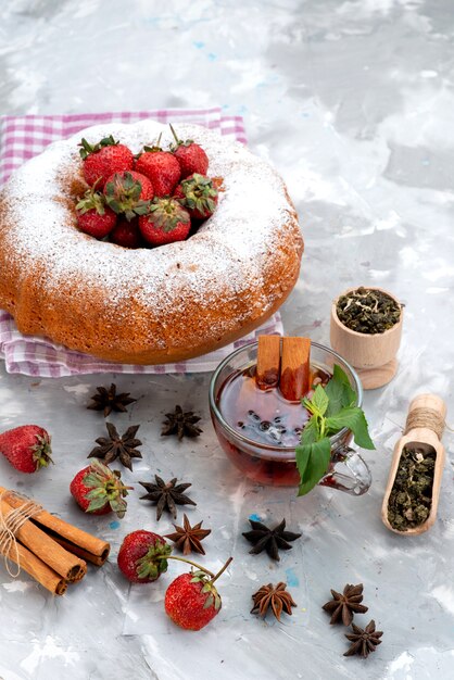 Una torta rotonda di vista superiore con tè alla cannella delle fragole rosse della polvere dello zucchero sulla torta bianca della frutta della bacca dello scrittorio