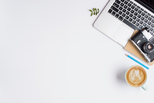 Una vecchia macchina fotografica; notebook sul portatile con matita blu e tazza di cappuccino su sfondo bianco