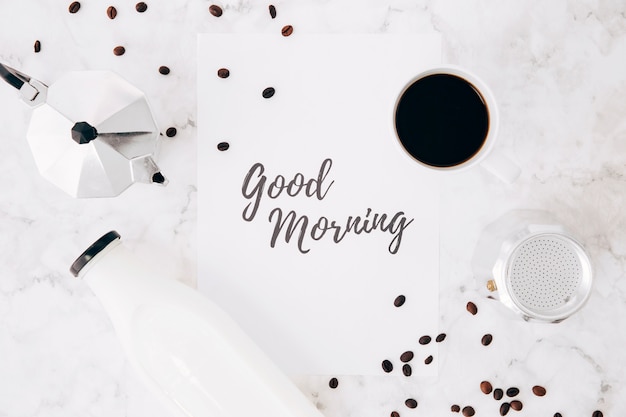 Una visione elevata del buongiorno testo su carta; caffettiera caffetteria; tazza di caffè; bottiglia di latte e chicchi di caffè sullo sfondo di marmo