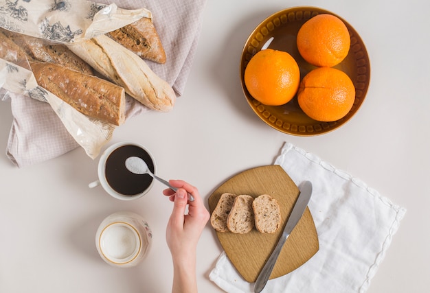 Una vista aerea della mano di una donna aggiungendo latte in polvere nella tazza di tè con pane e arance su sfondo bianco