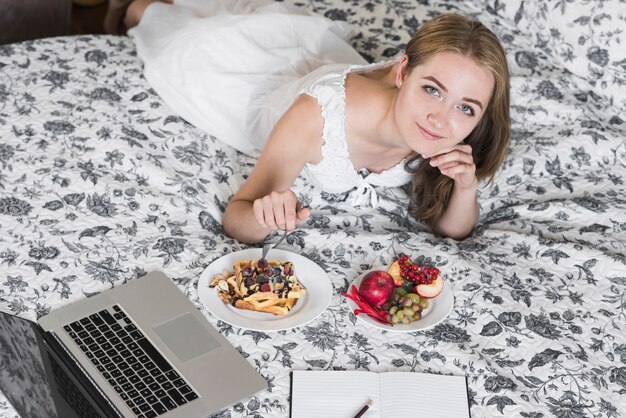 Una vista aerea di una giovane donna sdraiata sul letto inserendo la forchetta nella colazione waffle guardando la fotocamera