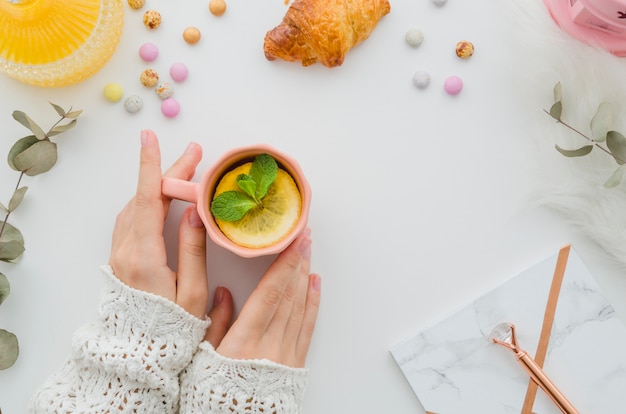 Una vista ambientale della donna che tiene tazza di tè al limone sul tavolo bianco con croissant e caramelle su priorità bassa bianca