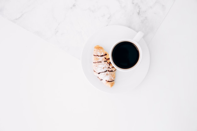 Una vista ambientale della tazza di caffè e del croissant sulla zolla bianca contro il contesto strutturato