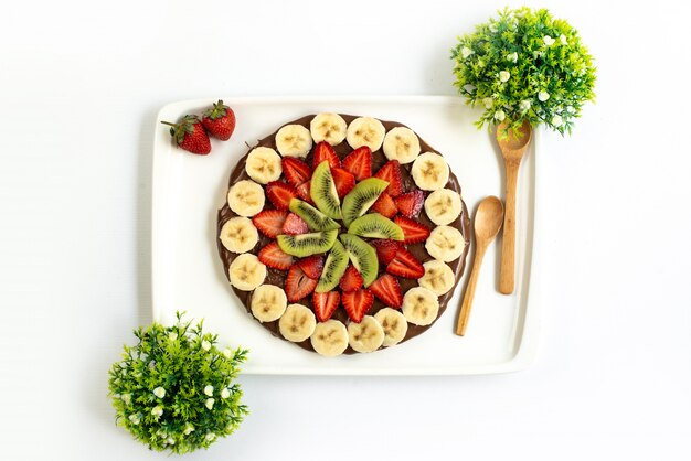 Una vista dall'alto choco fragola dessert gustoso dolce decorato banane fresche e kiwi insieme a piante sparse su tutto lo sfondo bianco torta di frutta esotica