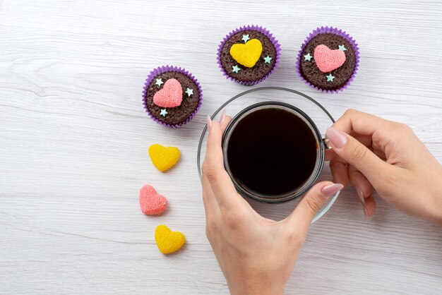 Una vista dall'alto deliziosi brownies all'interno di forme viola insieme a una tazza di tè tenuta da donna su dolci color bianco e caramelle