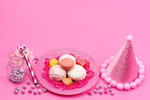 Una vista dall'alto meringhe e macarons dolci e deliziose torte all'interno della piastra con tappo di compleanno e fischietto di compleanno su rosa, biscotto cakek