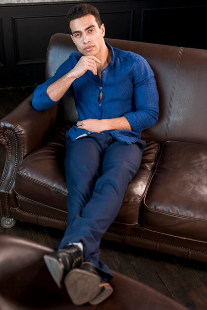 Una vista elevata del giovane bello che si siede sul sofà marrone con la mano sul suo mento che guarda l&#39;obbiettivo