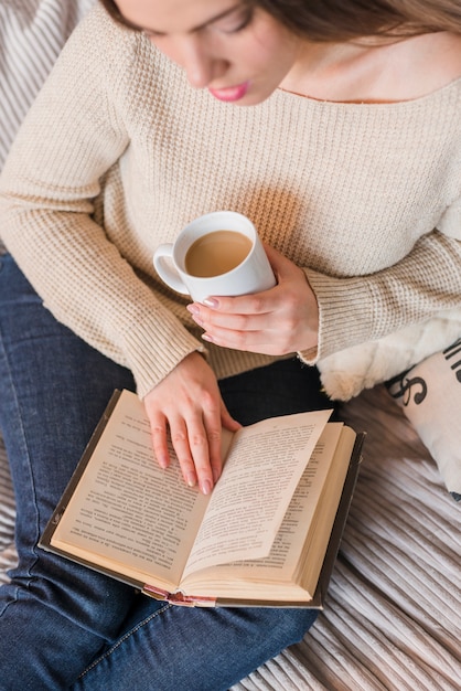 Una vista elevata del libro di lettura disponibila della tazza di caffè della tenuta della giovane donna