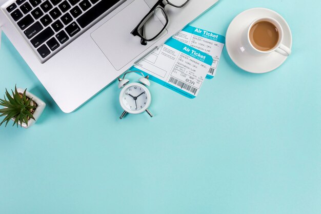 Una vista elevata di due biglietti aerei con tazza di caffè, laptop, occhiali da vista, sveglia sulla scrivania blu