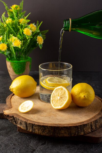 Una vista frontale acqua con limone fresca bevanda fresca che versa nel bicchiere con fette di limoni insieme a limoni interi e fiori sul buio