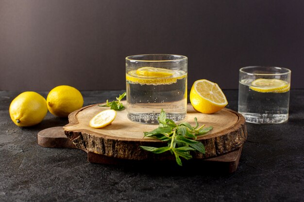 Una vista frontale acqua con limone fresca bevanda fresca con fette di limoni insieme a limoni interi e foglie all'interno di bicchieri trasparenti sul buio