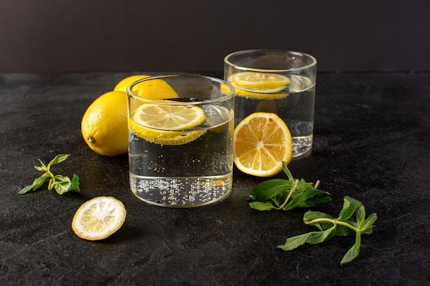 Una vista frontale acqua con limone fresca bevanda fresca con fette di limoni insieme a limoni interi e foglie all'interno di bicchieri trasparenti sul buio