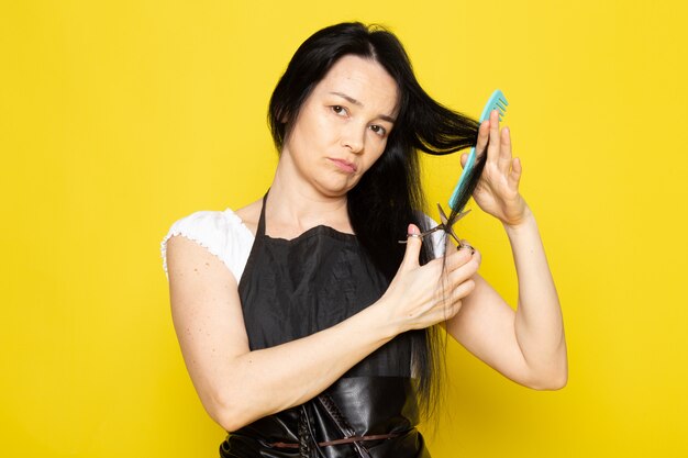 Una vista frontale bella femmina parrucchiere in maglietta bianca mantello nero con spazzole con capelli lavati spazzolatura e taglio dei capelli in posa sullo sfondo giallo stilista capelli da barbiere