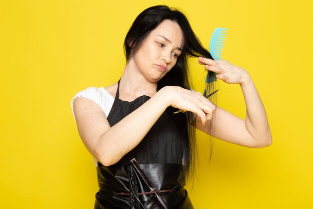Una vista frontale bella femmina parrucchiere in maglietta bianca mantello nero con spazzole con capelli lavati spazzolatura e taglio dei capelli in posa sullo sfondo giallo stilista capelli da barbiere