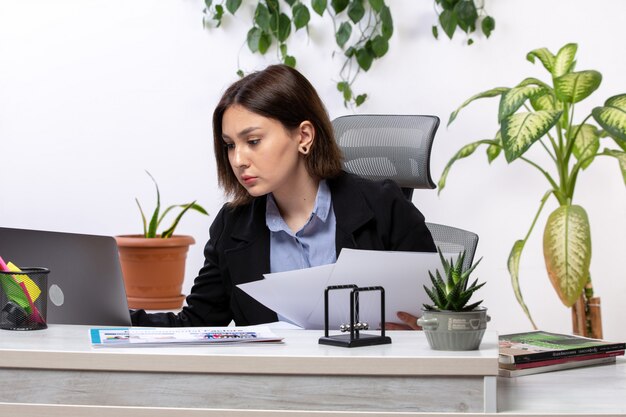 Una vista frontale bella giovane imprenditrice in giacca nera e camicia blu, lavorando con laptop e documenti di fronte al tavolo di lavoro ufficio di lavoro