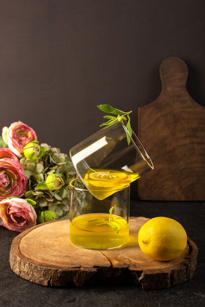 Una vista frontale bicchieri con succo di succo di limone all'interno di bicchieri trasparenti lungo tutto il limone e fiori sulla scrivania in legno marrone e sfondo grigio cocktail bevanda al limone