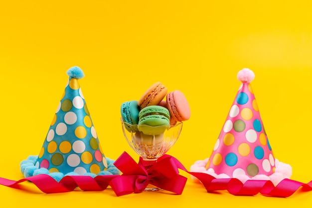 Una vista frontale bithday caps insieme a macarons francesi isolati su giallo, festa di compleanno di zucchero
