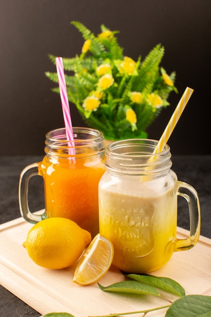 Una vista frontale cocktail freddi colorati all'interno di lattine di vetro con cannucce colorate limoni foglie verdi fiori sulla scrivania in legno crema e buio