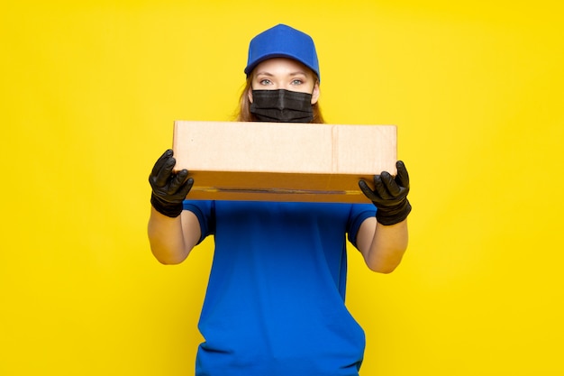 Una vista frontale femminile attraente corriere in polo blu berretto blu e jeans con pacchetto in guanti neri maschera protettiva nera su sfondo giallo lavoro di ristorazione