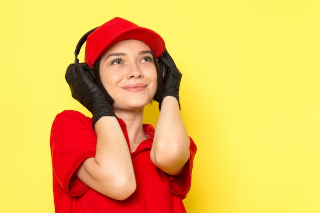 Una vista frontale giovane corriere femminile in uniforme rossa guanti neri e berretto rosso ascoltando musica con espressione felice