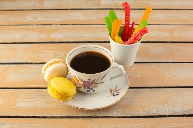 Una vista frontale tazza di caffè caldo e forte con macarons francesi e marmellata di arance sulla scrivania rustica color crema bere caffè foto forte