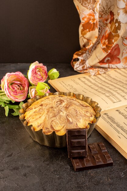 Una vista frontale torta rotonda dolce squisita deliziosa all'interno della tortiera insieme a choco bar fiori e note musicali quaderno sullo sfondo grigio biscotto di zucchero biscotto