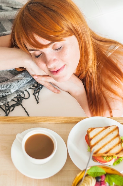 Una vista sopraelevata della donna sorridente che riposa vicino alla prima colazione sul vassoio di legno