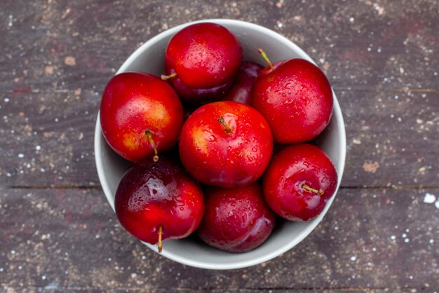Una vista superiore prugne rosse fresche pastose e mature all'interno del piatto bianco sul succo di polpa di frutta dello scrittorio di legno