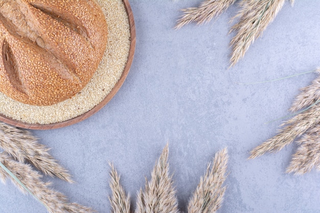 Unica pagnotta di pane in un vassoio pieno di semi di sesamo, circondata da gambi di erba piuma essiccata su una superficie di marmo