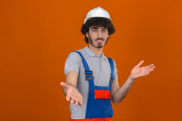 Uniforme d'uso della costruzione del giovane costruttore bello barbuto e casco di sicurezza che esaminano la macchina fotografica che sorride a braccia aperte per l'abbraccio sopra la parete arancio isolata