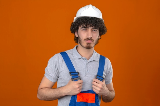 Uniforme d'uso della costruzione del giovane costruttore bello barbuto e casco di sicurezza che stanno con l'espressione di disapprovazione sulla persona negativa del fronte sopra la parete arancio isolata