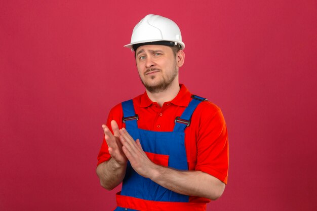 Uniforme da costruzione dell'uomo del costruttore e casco di sicurezza che applaudono con lo sguardo sicuro che controlla parete rosa isolata