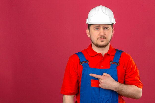 Uniforme da costruzione dell'uomo del costruttore e casco di sicurezza che indicano dito per copiare spazio con l'espressione triste sul fronte che controlla parete rosa isolata