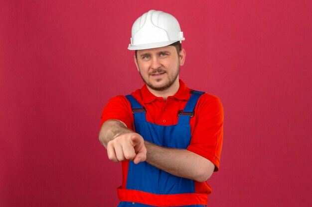 Uniforme da costruzione dell'uomo del costruttore e casco di sicurezza che sorridono e che esaminano la macchina fotografica che indica con il dito la macchina fotografica che controlla parete rosa isolata