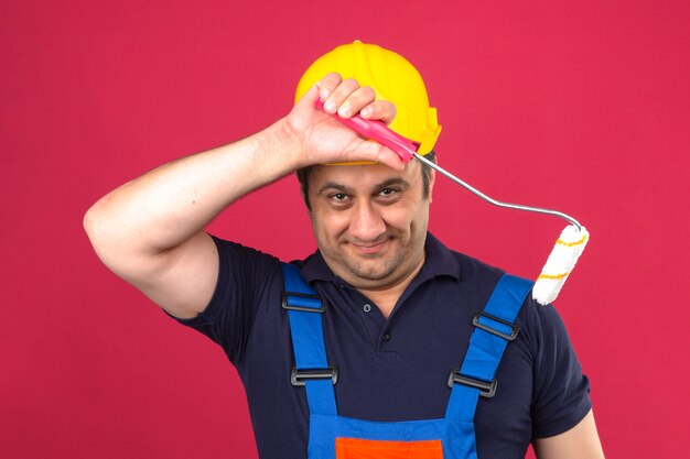 Uniforme da costruzione dell'uomo del costruttore e casco di sicurezza che stanno con il rullo di pittura che sembra felice e che sorride sopra la parete rosa isolata